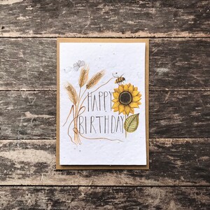 May include: A white card with a brown border features a hand-drawn illustration of a sunflower, wheat stalks, and a bee. The text "Happy Birthday" is written in black cursive.