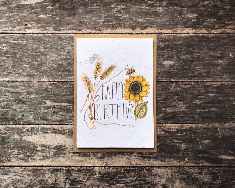 May include: A white card with a brown border features a hand-drawn illustration of a sunflower, wheat stalks, and a bee. The text "Happy Birthday" is written in black cursive.