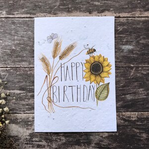 May include: A white card with a sunflower, wheat stalks, a bee, and the text "Happy Birthday" in black script.
