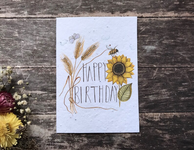 May include: A white card with a sunflower, wheat stalks, a bee, and the text "Happy Birthday" in black script.