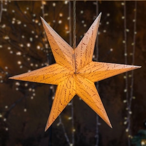 May include: A six-pointed star-shaped paper lantern, illuminated with a warm glow. The beige paper is printed with script, and the lantern hangs from a thin cord. The blurred background features soft lights, creating a festive atmosphere.