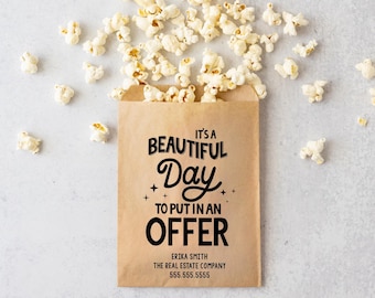 May include: A brown paper bag filled with popcorn with the text "It's a Beautiful Day to Put in an Offer" printed on the front. The bag also includes the name "Erika Smith", "The Real Estate Company", and a phone number.