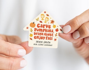 May include: A house-shaped real estate business card with a fall theme. It features illustrations of pumpkins, apples, leaves, and mugs of cider. The text reads "Carve Pumpkins, Drink Cider, Enjoy Fall" and "Erika Smith Real Estate Company".