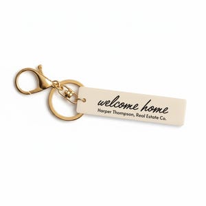 May include: A gold-colored keychain with a rectangular tag. The tag is cream-colored and features the words "welcome home" in a script font, along with the text "Harper Thompson, Real Estate Co."