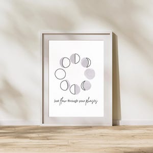Pode incluir: Uma impressão emoldurada branca com uma ilustração em preto e branco das fases da lua. O texto "Just flow through your phases" é impresso abaixo da ilustração.