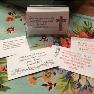 May include: A clear acrylic card holder containing a stack of white rectangular cards with printed religious text and colorful cross graphics. Several individual cards are displayed in front, featuring various Bible verses addressed to Katie.