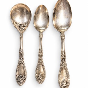 May include: Three silver-toned metal spoons with ornate embossed detailing on the handles and bowls. The handles feature intricate grape cluster motifs and vine patterns, with a polished finish showing signs of natural metal oxidation.