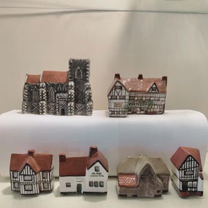 May include: A collection of six miniature ceramic buildings featuring a grey stone church, a timber-framed house, a cottage, a white building labeled Six Bells Greene King, a barn with a brown roof, and a small black and white timber-framed house.