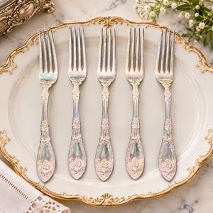 May include: A set of five silver-toned metal forks with ornate, embossed floral detailing on the handles. The forks are arranged in a parallel row on a cream-colored ceramic plate featuring a scalloped edge with gold-painted trim.