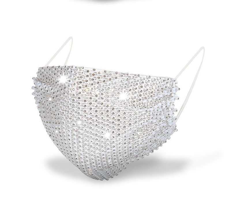 May include: A white face mask covered in rhinestones. The mask has a simple design with two straps that tie behind the head.