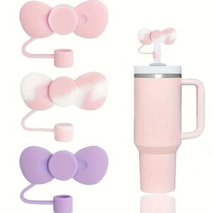 May include: Three silicone bow-shaped bottle stoppers with a loop handle. The stoppers are in pink and purple. The stoppers are shown against a white background.