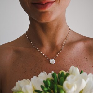 Può includere: Una donna indossa una delicata collana di perle con un grande ciondolo di perla bianca irregolare. Sta tenendo un mazzo di fiori bianchi.