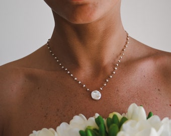 May include: A woman wearing a delicate pearl necklace with a large, white, irregular pearl pendant. She is holding a bouquet of white flowers.