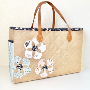 May include: A brown tote bag with a quilted design and three fabric flower appliques. The bag has brown leather handles and a black trim.