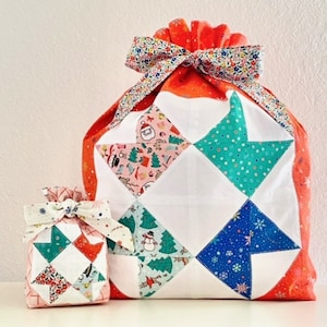 May include: Two red and white fabric gift bags with a patchwork design. The larger bag has a white bow with a floral pattern. The smaller bag has a white bow with a red and white floral pattern.