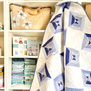 May include: A white quilt with a blue and white geometric pattern. The quilt is folded and draped over a shelf with books and fabric scraps.