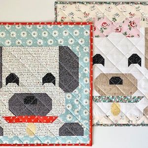 May include: Two quilted dog-shaped wall hangings. The dog on the left is gray with a red collar and a gold tag. The dog on the right is white with a brown nose and a green collar. Both dogs have black eyes and are surrounded by a patterned fabric border.