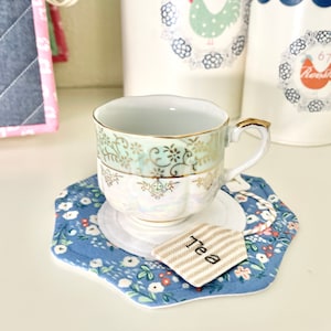 May include: A vintage teacup and saucer with a floral pattern and gold trim. The saucer is blue with a white center and a floral pattern. A small tag with the word "tea" is on the saucer.