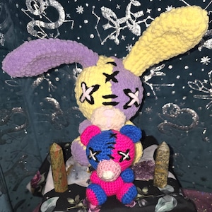 May include: Two crocheted plush toys, a purple and yellow bunny and a pink and blue bear, both with stitched-on black X's for eyes. The toys are sitting on a black surface with green and purple crystals.