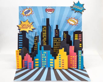 Könnte beinhalten: Eine Pop-up-Karte mit einer Stadtlandschaft mit bunten Gebäuden und Sprechblasen im Comic-Stil, die "POW!" "BOOM!" "ZAP!" und "BOOM!" sagen.