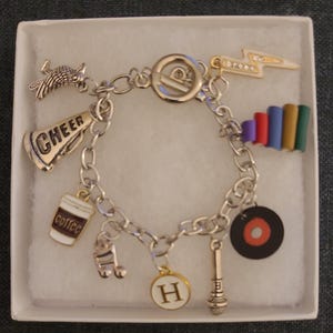 May include: A silver charm bracelet with various charms, including a megaphone with "CHEER", a coffee cup, a musical note, a letter "H", a microphone, a record, a lightning bolt, and a stack of books. The bracelet is displayed in a white box.