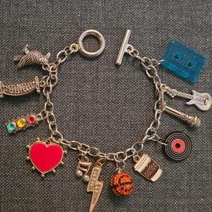 May include: A silver charm bracelet with a variety of charms, including a heart, a coffee cup, a basketball, a lightning bolt, a traffic light, a record, a microphone, a guitar, a cassette tape, a bird, and a tower.