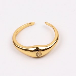 Puede incluir: Un anillo dorado con una banda redondeada ligeramente curvada. El anillo tiene un pequeño diseño grabado en la parte superior.