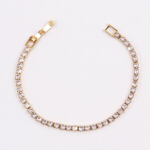 Może przedstawiać: Złota bransoletka tenisowa z rzędem przezroczystych, okrągłych kamieni szlachetnych. Bransoletka jest pokazana na białym tle, z otwartym zapięciem. Kamienie szlachetne są równomiernie rozmieszczone i odbijają światło.