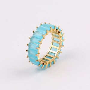 Può includere: Un anello d'oro con una fila di pietre rettangolari di colore azzurro chiaro. Le pietre sono incastonate in una fascia d'oro e hanno un bordo leggermente appuntito.