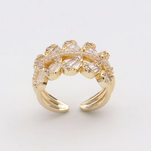 Könnte beinhalten: Ein goldener Ring mit einem filigranen Design, das einen Cluster aus klaren Zirkonia-Steinen zeigt. Der Ring hat ein breites Band und ein einzigartiges, durchbrochenes Design.