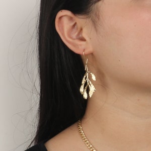 Puede incluir: Pendientes de hoja de oro con un delicado diseño de rama. Los pendientes se muestran en la oreja de una modelo.