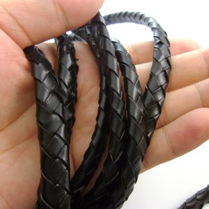 Puede incluir: Cordón de cuero trenzado negro, de aproximadamente 1,2 cm de ancho, en una mano. El cordón está hecho de múltiples hebras de cuero que se tejen juntas para crear un diseño duradero y elegante.