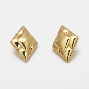 Puede incluir: Un par de pendientes de botón dorados geométricos. Cada pendiente presenta un diseño en forma de diamante en capas con una superficie pulida y reflectante. Los pendientes están sobre un fondo blanco.