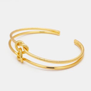 May include: A gold-colored cuff bracelet with a knot design. The bracelet features a double-band design, with one band having a textured rope-like pattern. The knot is centered, adding a unique focal point to the jewelry.