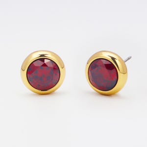 May include: A pair of stud earrings with a gold-colored bezel and a faceted, deep red stone. The earrings are round and have a post for pierced ears. The red stone has a speckled appearance.