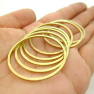 May include: A collection of thin, gold-colored metal rings, oval in shape, are arranged in a hand. The rings are stacked together, creating a layered effect. The background is a soft, neutral tone, highlighting the metallic sheen of the rings.