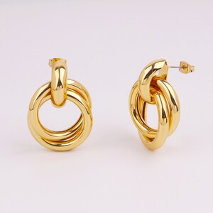 May include: A pair of gold-colored earrings. Each earring features a large, circular design with multiple interlocking rings. The earrings have a shiny, polished finish and a post-back closure. The jewelry is displayed against a plain white background.