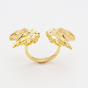 Può includere: Un anello color oro con un design diviso. L'anello presenta due elementi dettagliati a forma di ala su entrambi i lati della divisione. Le ali hanno superfici strutturate e sono posizionate per creare un design aperto. L'anello è posto su uno sfondo bianco.
