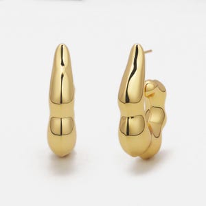 May include: Two gold-colored hoop earrings. The earrings have a unique, organic shape with a smooth, reflective surface. The design features a series of rounded, bulbous forms, creating a modern and sculptural aesthetic. The earrings are displayed against a white background.