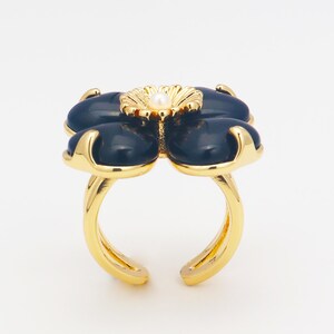 Könnte beinhalten: Goldfarbener Ring mit einem dunkelblauen Blumenmuster. Die Blume hat vier Blütenblätter und eine goldene Mitte mit einer kleinen Perle. Der Ring ist unten offen.