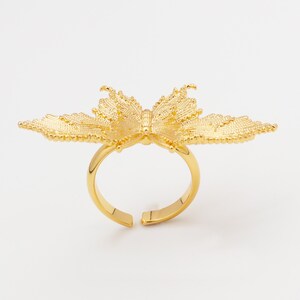 Könnte beinhalten: Ein goldfarbener Schmetterlingsring mit filigranen Details. Die Schmetterlingsflügel erstrecken sich von der Ringschiene aus und bilden ein einzigartiges Design. Der Ring ist verstellbar und vor weißem Hintergrund.