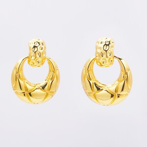 May include: A pair of gold-coloured earrings. Each earring has a textured, rectangular top piece and a large, quilted, circular drop. The earrings have a shiny, reflective surface and a classic, elegant design.