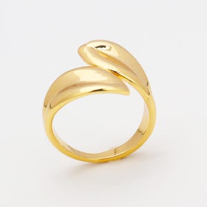 Könnte beinhalten: Ein goldfarbener Ring mit einem einzigartigen, fließenden Design. Der Ring hat zwei geschwungene, blattartige Formen, die sich überlappen und einen offenen, verstellbaren Stil schaffen. Die polierte Oberfläche reflektiert das Licht und unterstreicht die glatte, elegante Form des Rings. Ein modernes Schmuckstück.