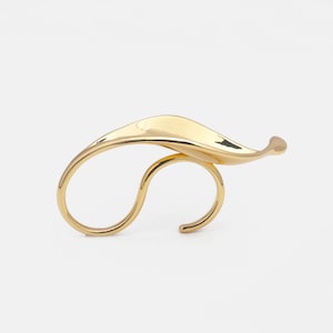 May include: A gold-toned ring with a unique, abstract design. The ring is made of a single piece of metal that is bent and twisted to create a flowing, organic shape.