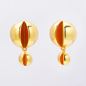 Pode incluir: Um par de brincos dourados. Cada brinco apresenta uma esfera grande e redonda de ouro polido com um detalhe vertical laranja-avermelhado. Uma esfera dourada menor pende abaixo, também com um detalhe laranja-avermelhado. Fundo branco.