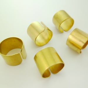 May include: Five gold-colored metal rings, each with a wide band and a slightly open top. The rings are arranged in a semi-circle on a white surface.