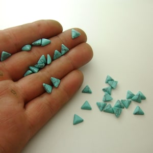 May include: A collection of turquoise triangle-shaped beads. The beads are scattered on a white surface and in a hand.