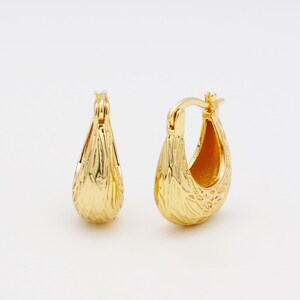 Peut inclure: Une paire de boucles d'oreilles créoles dorées. Les boucles d'oreilles ont une forme de goutte d'eau texturée avec un motif floral détaillé. Les boucles d'oreilles sont présentées sur fond blanc.