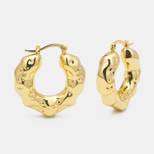 Pode incluir: Brincos de argola dourados com uma superfície texturizada irregular. Os brincos têm um design simples e moderno.