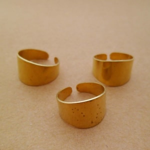 Könnte beinhalten: Drei goldfarbene Metallringe, jeder mit einem einfachen, flachen, rechteckigen Design. Die Ringe sind in einem dreieckigen Muster auf einer hellbraunen Oberfläche angeordnet.